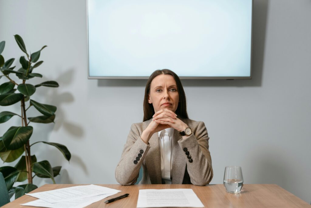 Businesswoman in a brown blazer seated, serious expression, office setting.